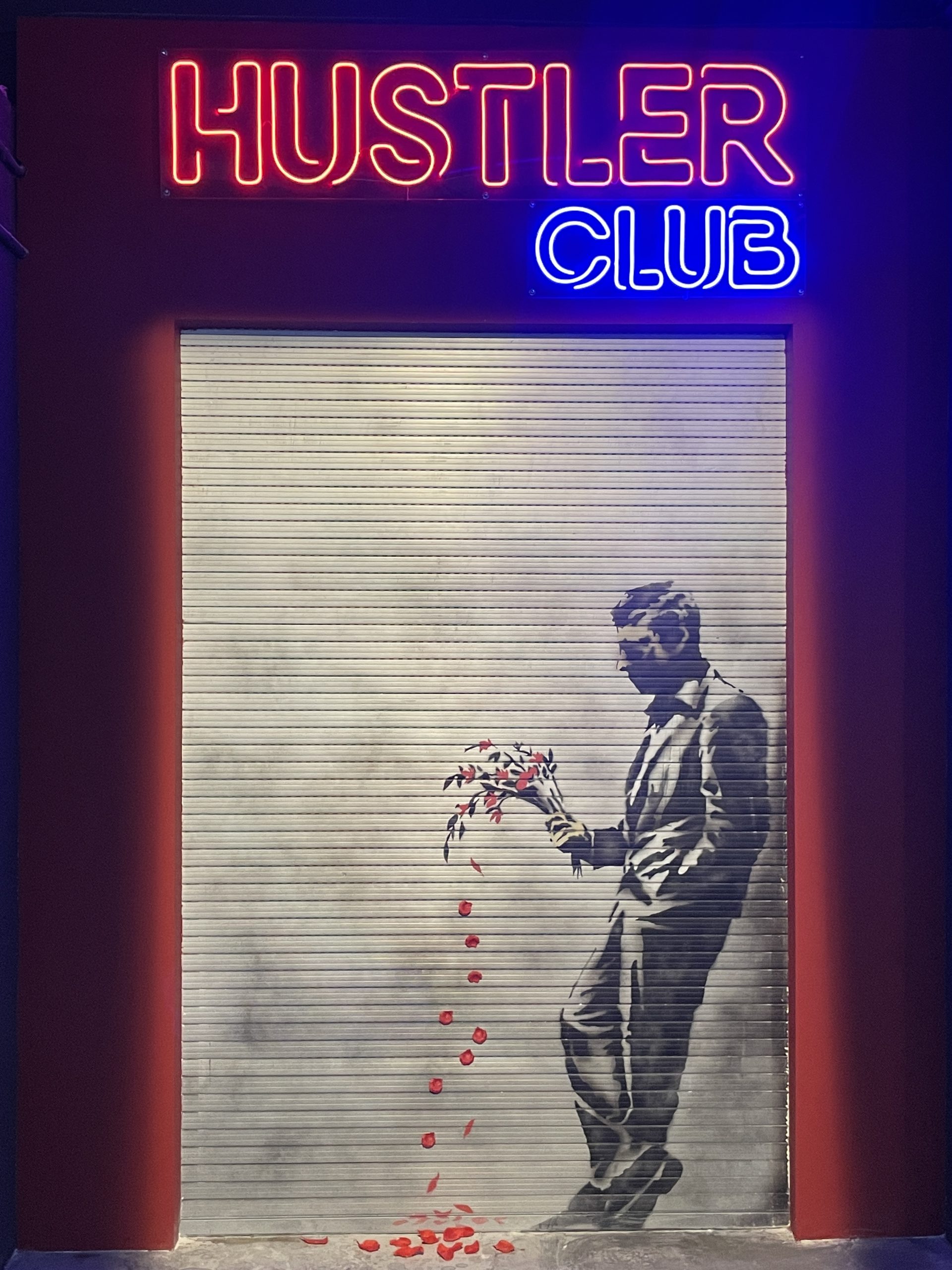 Letrero de neón 'HUSTLER CLUB' sobre una persiana metálica con una plantilla de un hombre sosteniendo flores, cayendo pétalos, iluminación roja y azul