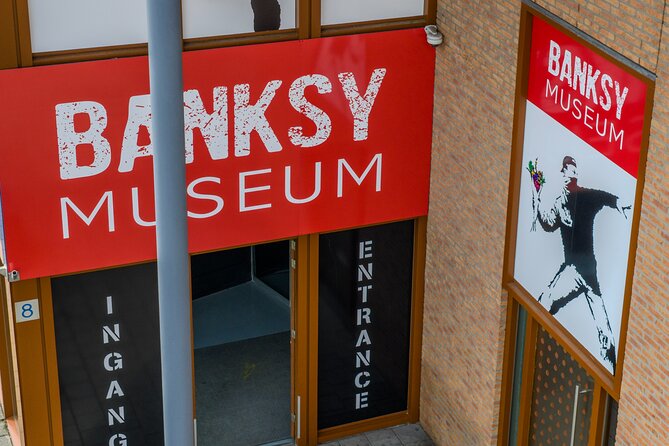 Cartel rojo con texto 'BANKSY MUSEUM' sobre las puertas de entrada con rotulación vertical 'INGANG' y 'ENTRANCE', edificio de ladrillo con obra de arte estilo Banksy