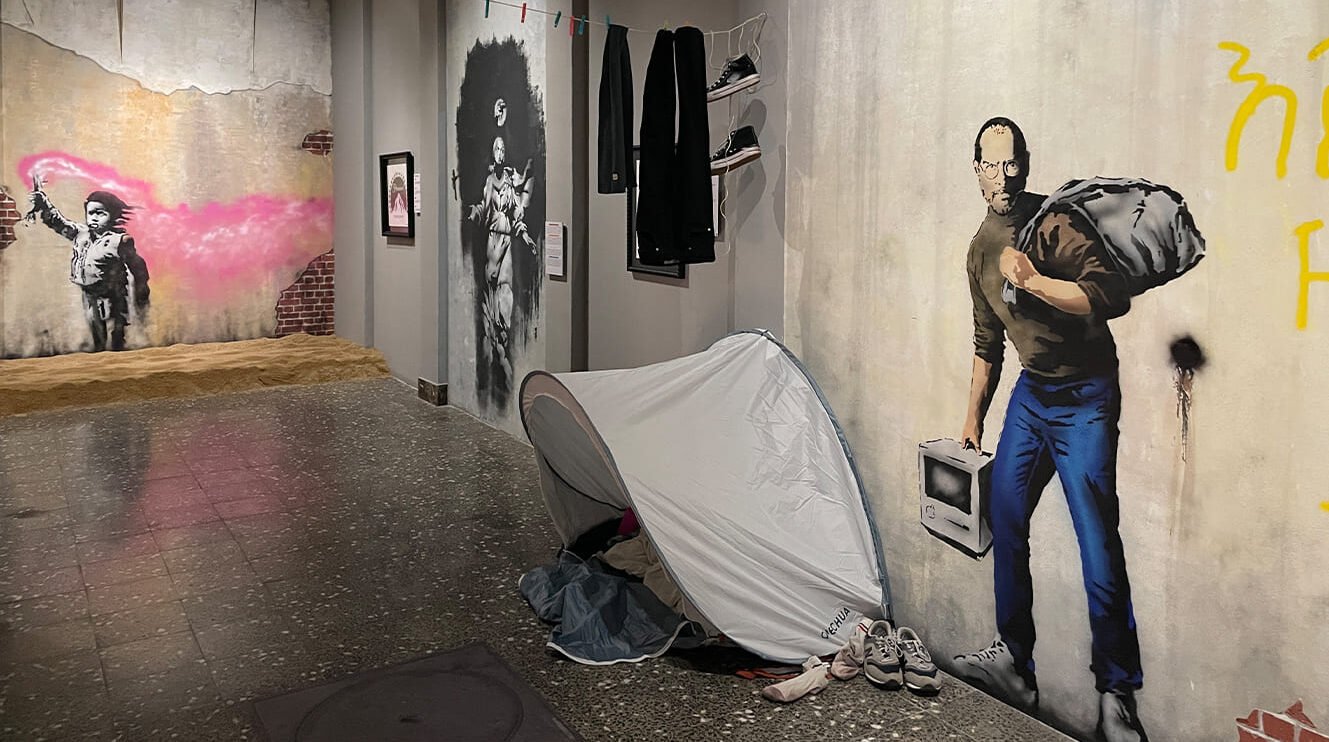 Interior del Museo Banksy de Ámsterdam que muestra murales de arte callejero, incluyendo una figura con spray, un retrato stencilado y un hombre cargando una bolsa con un televisor.