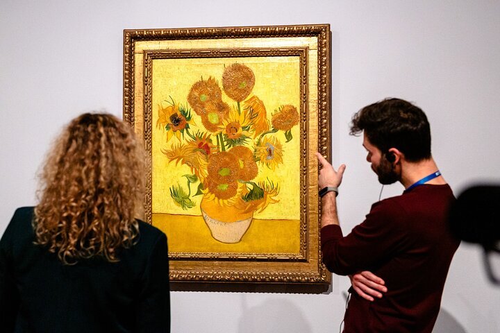 Dos personas observan un cuadro enmarcado de girasoles con pétalos dañados sobre una pared blanca, expuesto en un museo.