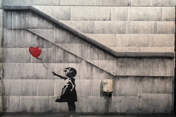 Black stencil mural of a girl reaching for a red heart-shaped balloon on a stone wall with 'THERE IS ALWAYS HOPE' text