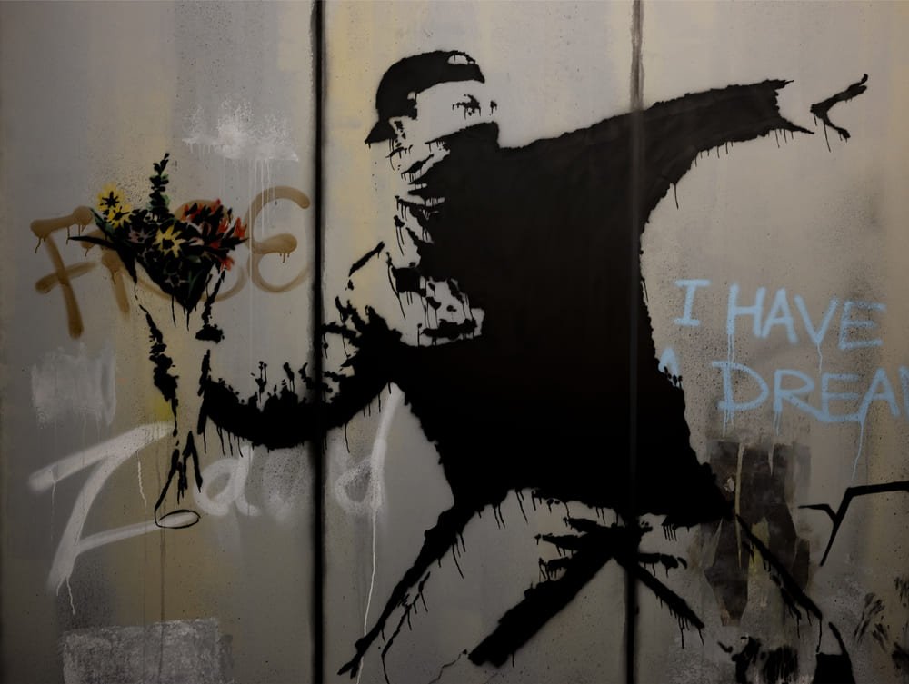 Silueta negra estarcida de una figura enmascarada lanzando un ramo de flores, con el texto 'I HAVE A DREAM' en una pared desgastada