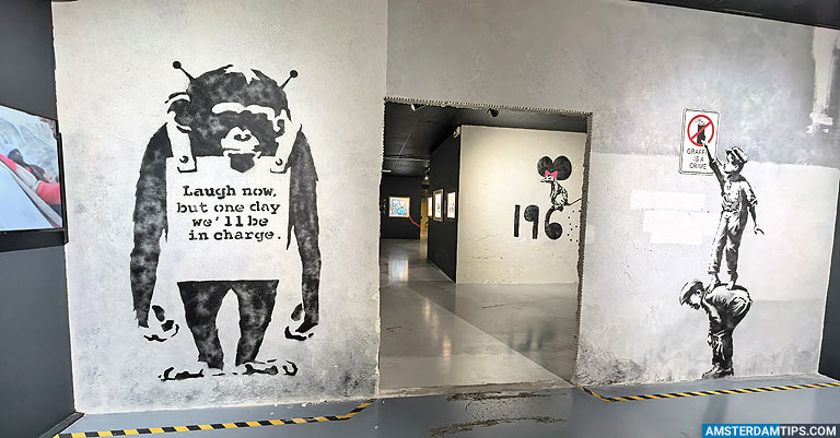 Pasillo de paredes de hormigón que exhibe murales estilo Banksy, que incluyen un mono con un cartel, una figura pintando a otra y un letrero de '196'