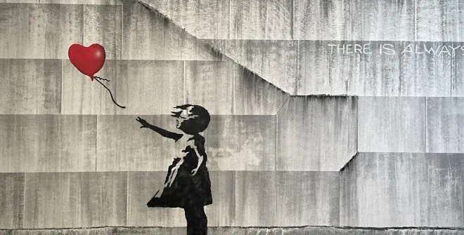 Un esténcil en blanco y negro de una niña que alcanza un globo rojo en forma de corazón en una pared de concreto con el texto 'THERE IS ALWAYS' al fondo.