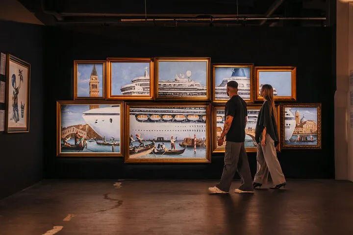 Twee mensen lopen door een galerie met grote ingelijste kunstwerken van schepen en stadsgezichten op donkere muren