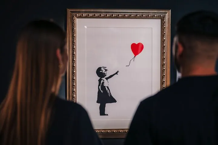 Een ingelijst kunstwerk van een meisje dat naar een rood hartvormig ballonnetje reikt, gezien van achter twee mensen in een galerie.