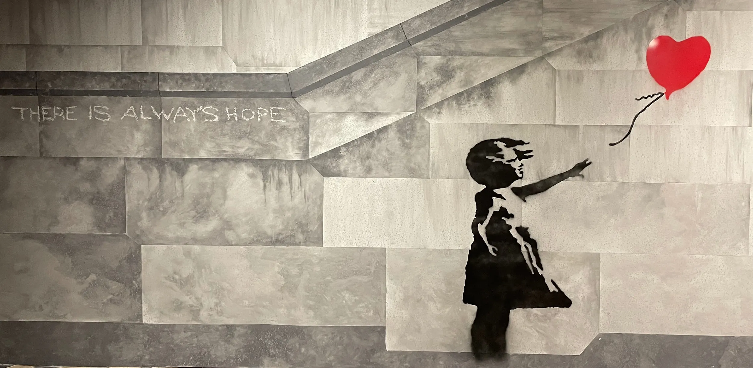 Mural estarcido de una niña que alcanza un globo rojo en forma de corazón en una pared gris con el texto 'SIEMPRE HAY ESPERANZA' arriba