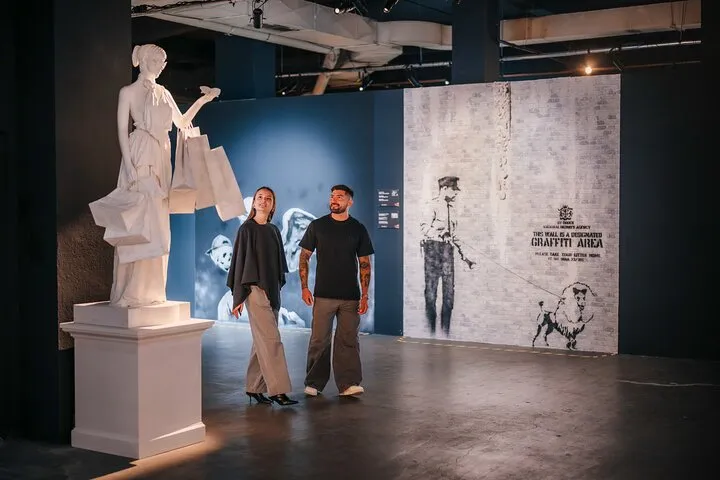 Twee mensen die in een tentoonstellingsruimte staan met een wit beeld op een sokkel en een grote muurschildering aan de muur met straatkunstfiguren en tekst.