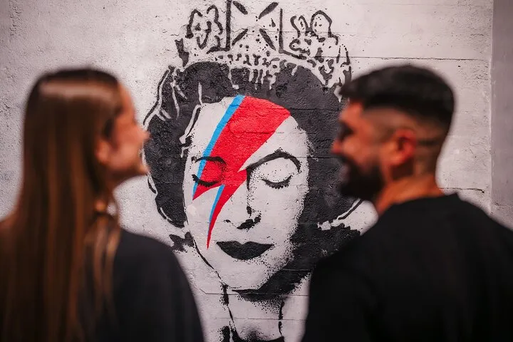 Dos personas observando un gran mural de la cara de una mujer con un rayo rojo, que recuerda a Ziggy Stardust de David Bowie, sobre una pared blanca.