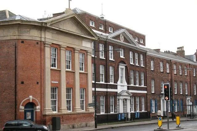 Red brick historic building facade with classical columns and triangular pediment, street view with traffic lights and parked cars