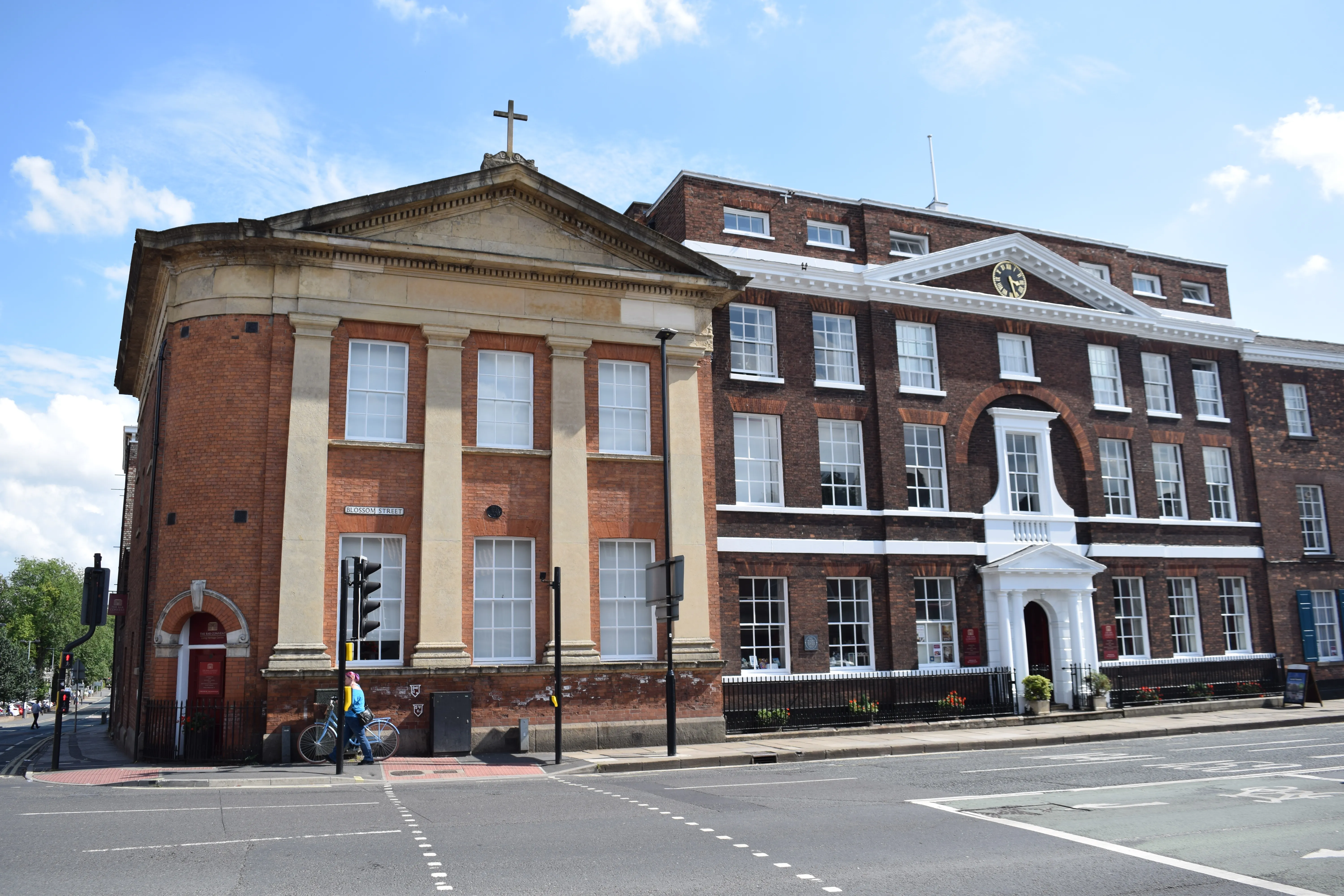 Exterior view of the Bar Convent Living Heritage Centre, a large red brick building with a classical portico facade, white entrance canopy, and a cross atop the roofline, viewed from across a street on a sunny day