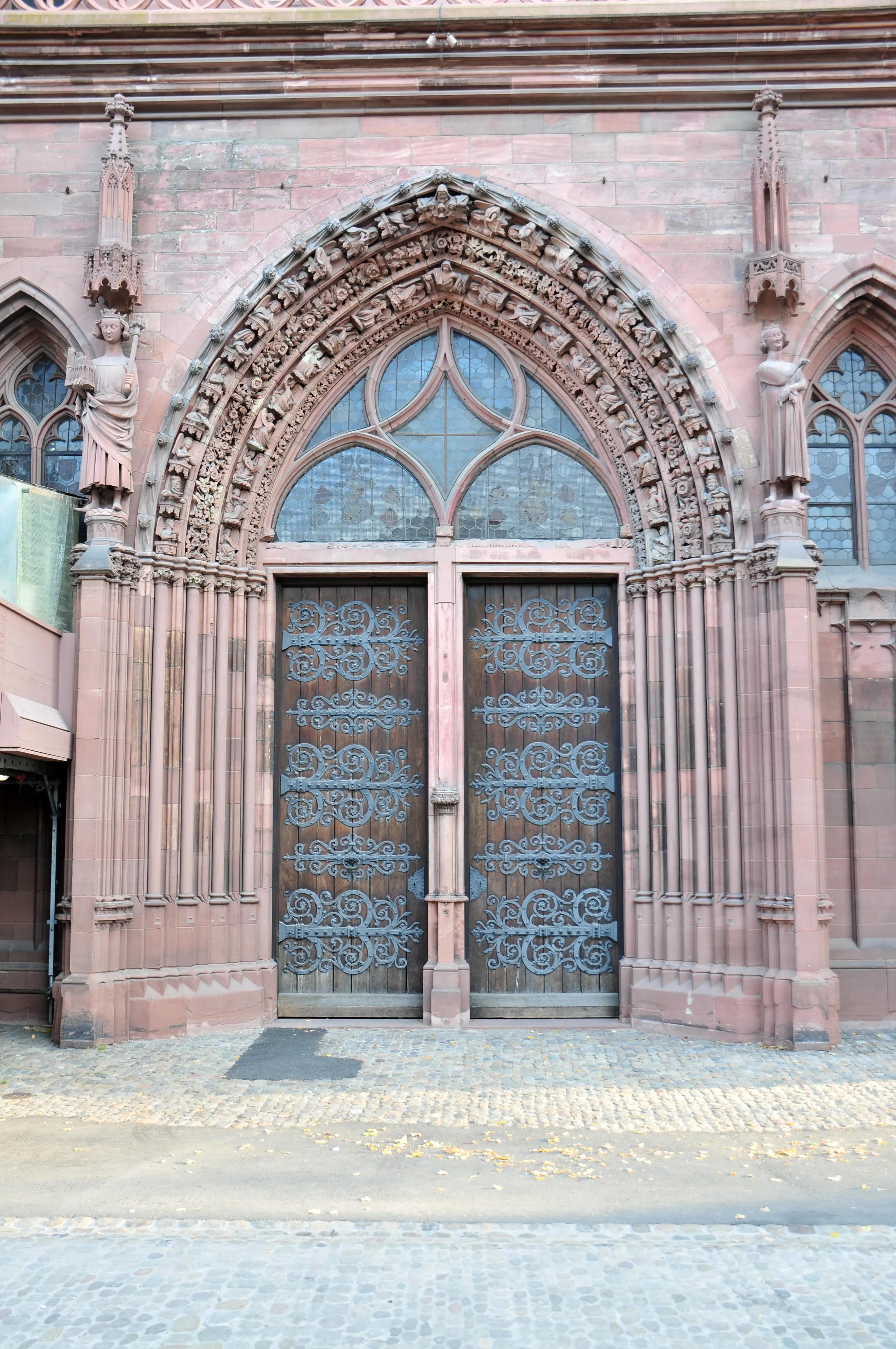 Ozdobny gotycki portal katedry Basel Minster z łukiem z czerwonego piaskowca, misternymi rzeźbieniami i kunszytymi elementami z kutego żelaza na drewnianych dwuskrzydłowych drzwiach.