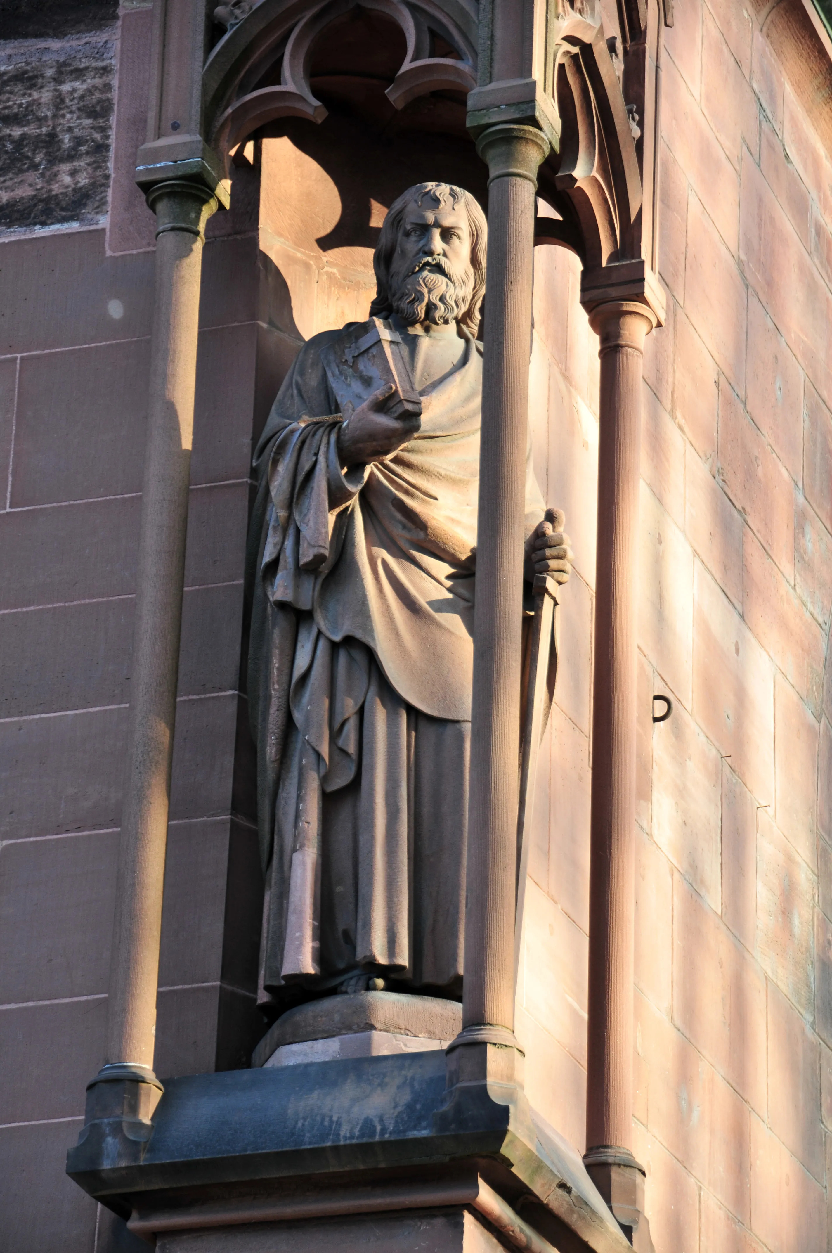 Kamienny posąg brodatej postaci w szatach trzymającej książkę, umieszczony w gotyckiej niszy architektonicznej na fasadzie budynku z czerwonego piaskowca.