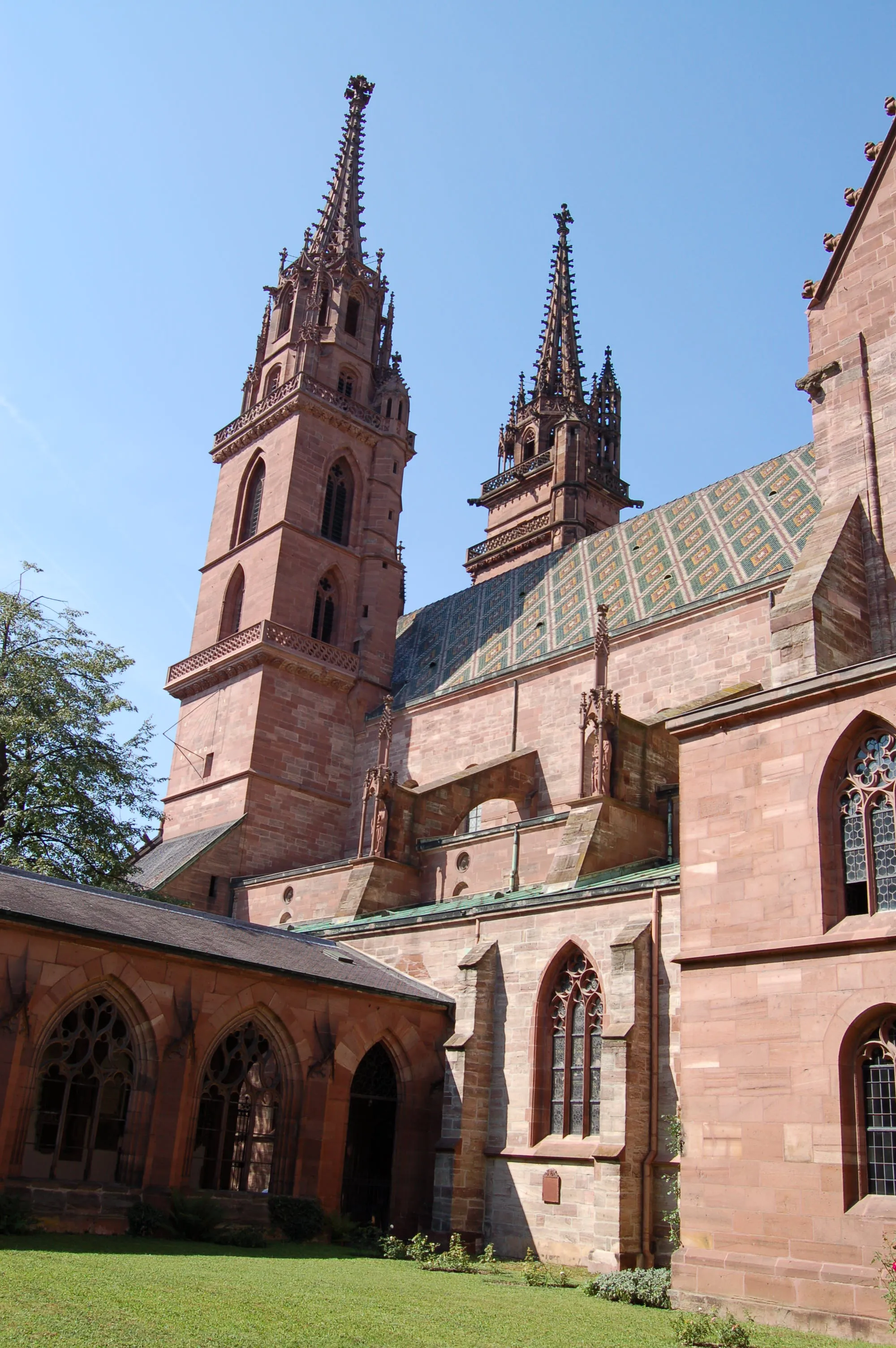 Widok z zewnątrz na katedrę Basler Münster, ukazujący bliźniacze wieże z czerwonego piaskowca, wzorzysty dach oraz przyległy krużganek z łukowymi oknami.