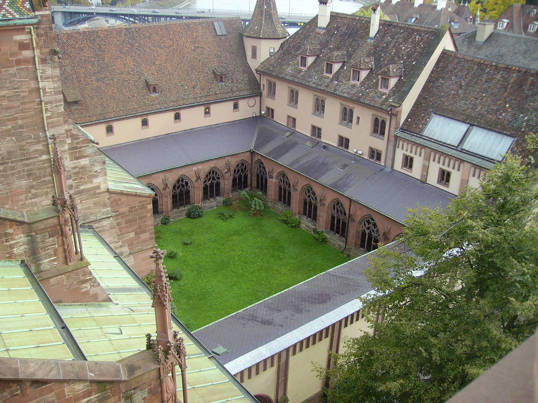 Widok z góry na dziedziniec krużganków katedry w Bazylei z zielonym trawnikiem otoczonym gotyckimi arkadami i architekturą z czerwonego piaskowca.