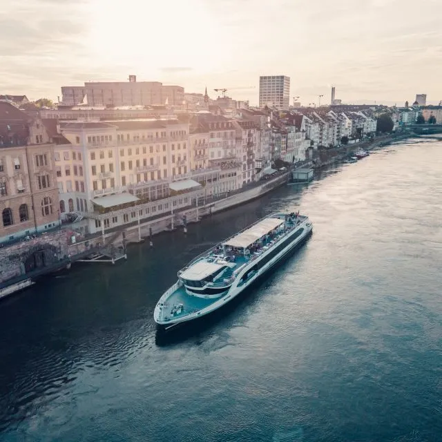 Luchtfoto van een lang wit en blauw cruiseschip dat over de Rijn vaart langs de historische panden aan de kade van Basel.