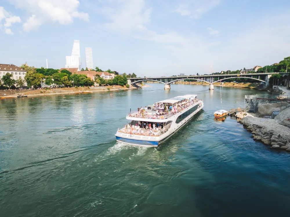 Een wit en blauw passagiersschip dat op de Rijn vaart met een stenen boogbrug en het stadsgezicht van Basel op de achtergrond onder een halfbewolkte lucht.