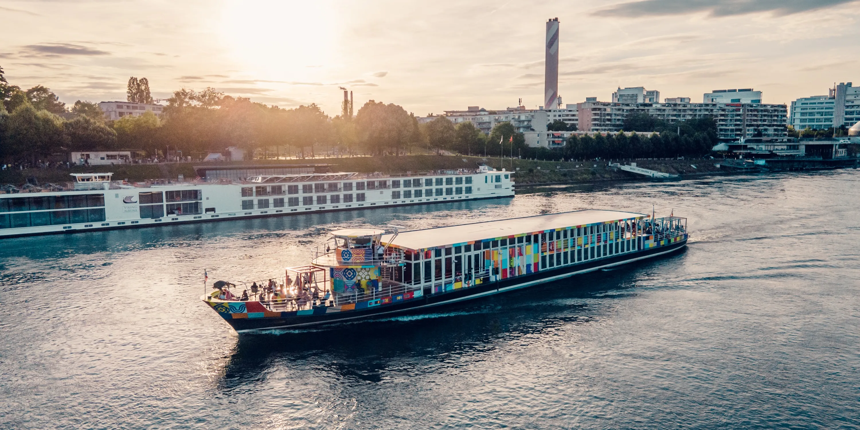 Een kleurrijke rondvaartboot vaart bij zonsondergang over de Rijn in Basel, met op de achtergrond een groter wit cruiseschip en de gebouwen van de stad.