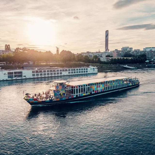 Een kleurrijke passagiersboot vaart bij zonsondergang op de Rijn, met de skyline van Basel en een hoge toren op de achtergrond zichtbaar.