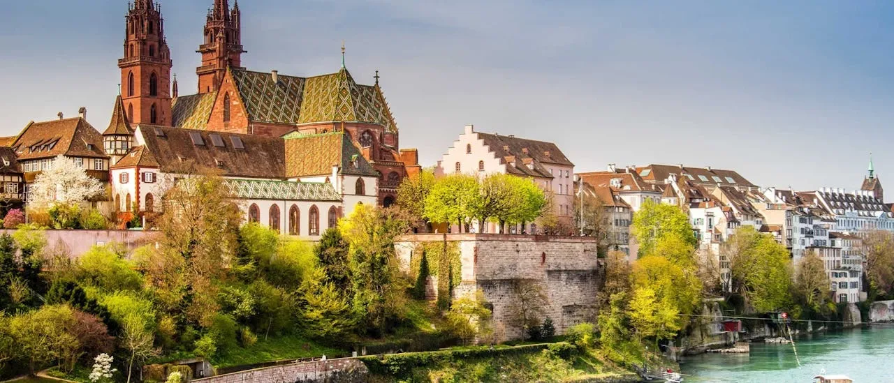 Panoramisch uitzicht op de oude binnenstad van Basel langs de Rijn, met de rode zandstenen Basel Minster-kathedraal met zijn karakteristieke kleurrijke pannendak, omringd door historische gebouwen en weelderige groene bomen.
