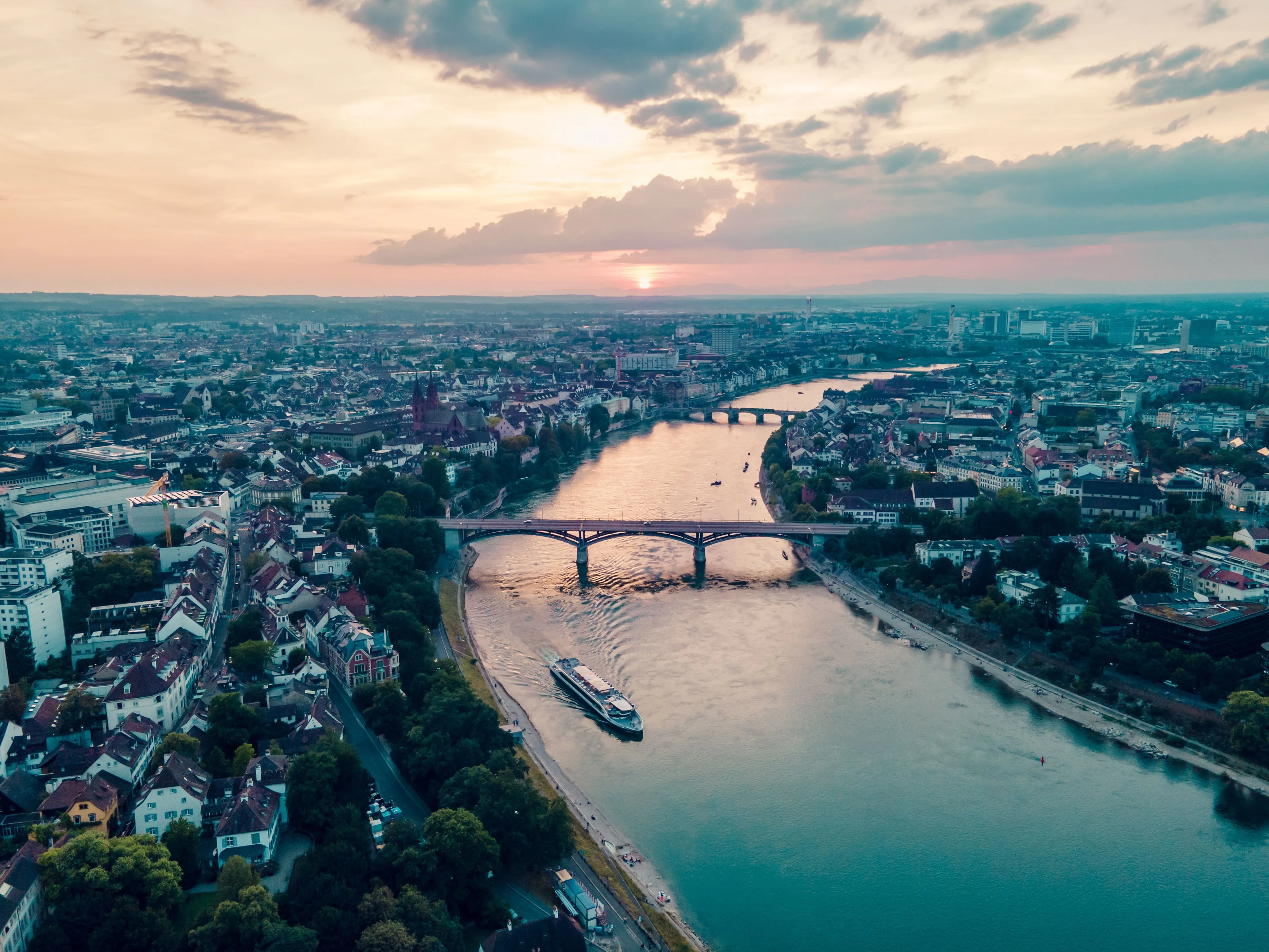 Luchtfoto van de rivier de Rijn die bij zonsondergang door Basel slingert, met een cruiseschip op het water en bruggen die de rivier overspannen.
