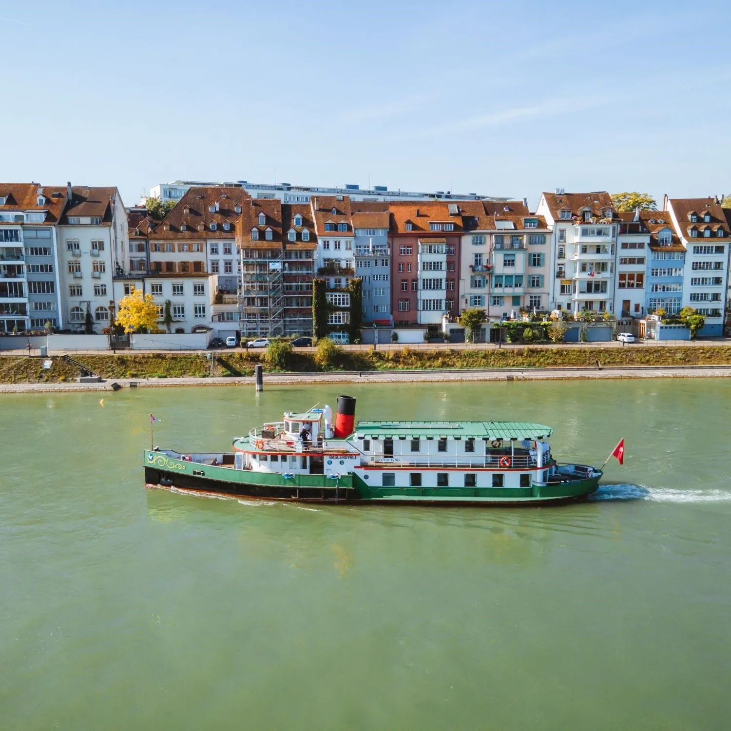 Een groen-witte riviercruiseschip varend op de Rijn met kleurrijke historische gebouwen van Basel langs de oever op de achtergrond.
