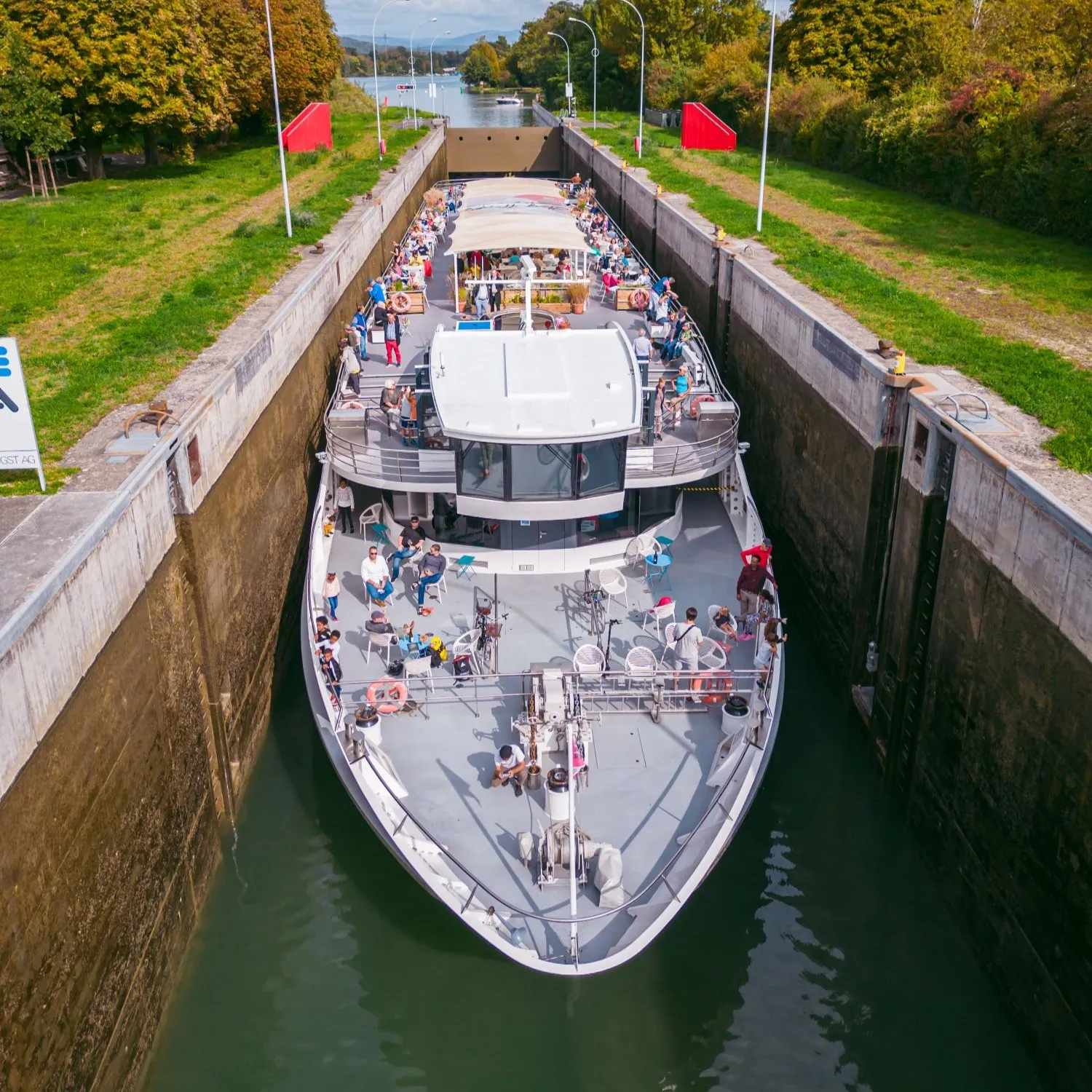 Luchtopname van een witte rondvaartboot vol passagiers die door een betonnen sluis op de Rijn vaart, geflankeerd door groene grasoevers en rode vlaggen.