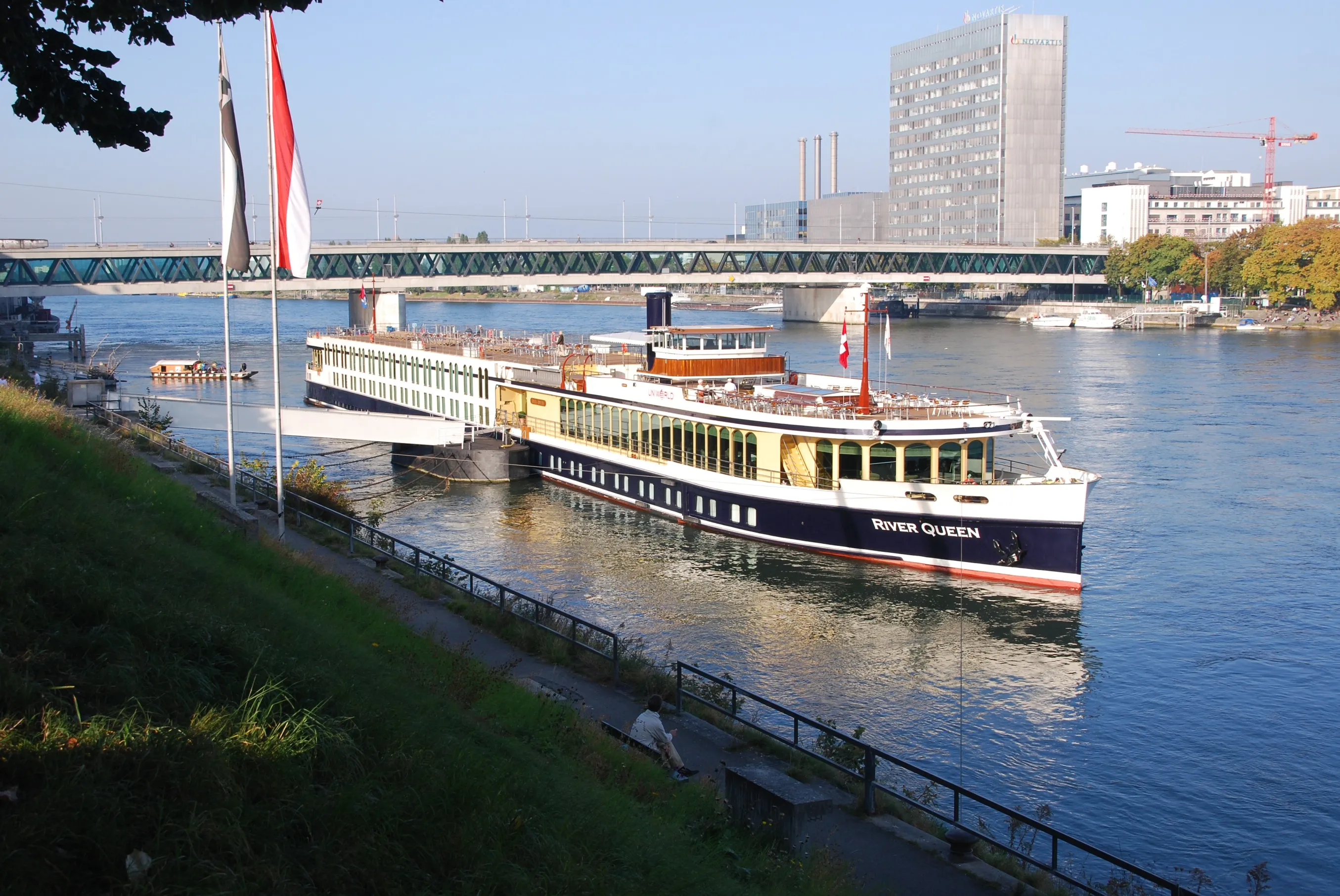 Een wit-blauwe riviercruiseschip genaamd Basel-Zürich aangemeerd op de Rijn met een groene vakwerkbrug en moderne gebouwen op de achtergrond