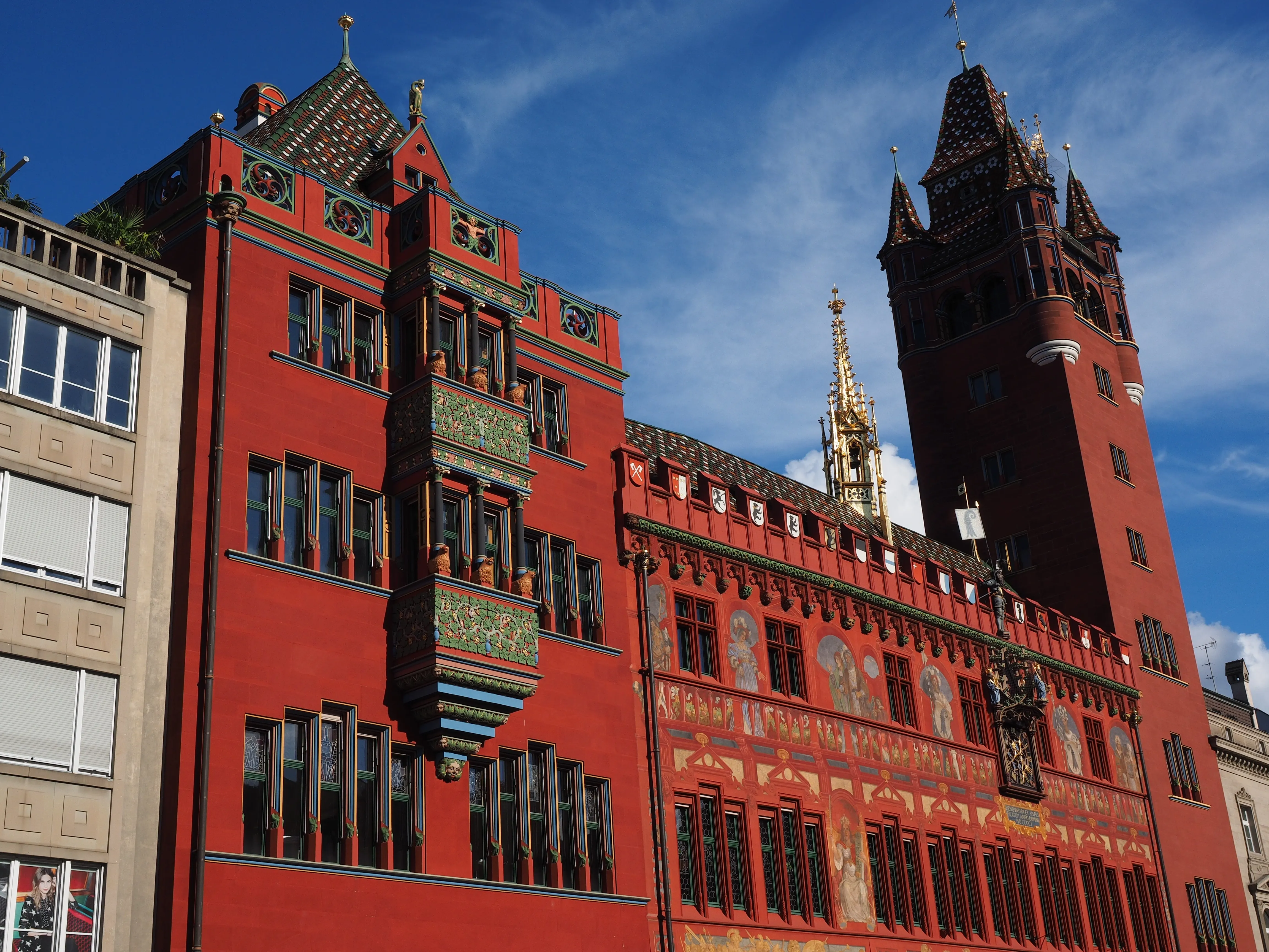 Levendig rood stadhuis van Bazel met een hoge donkere toren, beschilderde gevelpanelen, een sierlijke daklijn en een blauwe lucht op de achtergrond