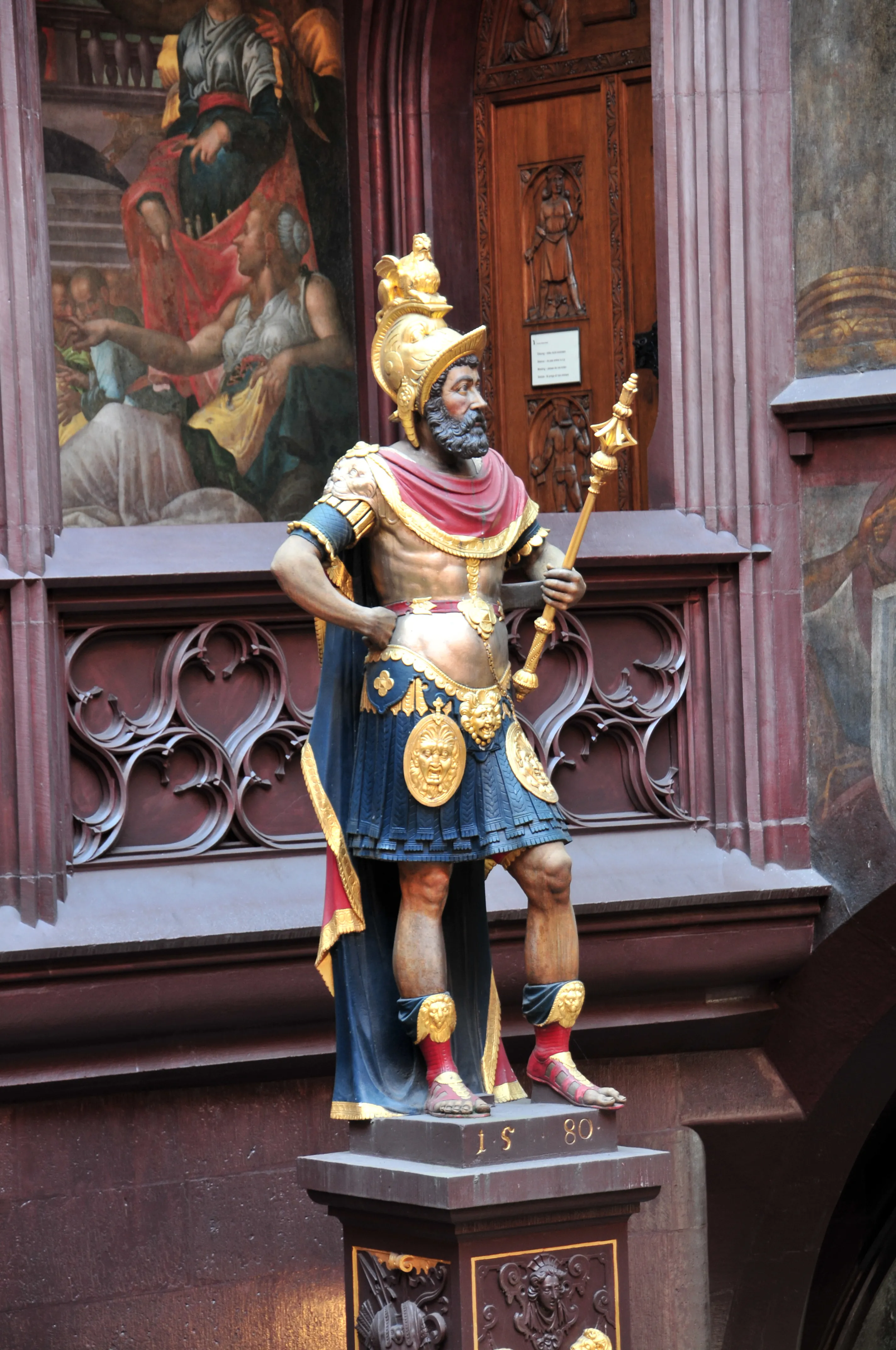 Een kleurrijk beschilderd standbeeld van een krijger met baard in Romeins aandoend harnas, met een gouden helm, blauwe tuniek met gouden medaillon en een rode cape; hij houdt een staf vast en staat op een sokkel met het jaartal 1580, voor een muur met fresco's.