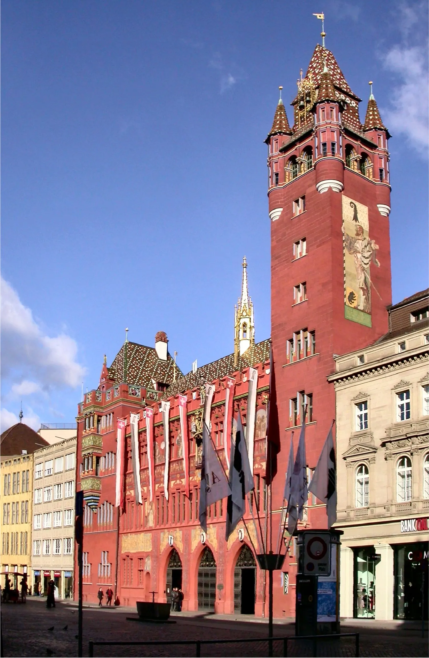 Rood stadhuis van Bazel met een hoge toren, kleurrijk pannendak, wapperende vlaggen en muurschilderingen op de gevel aan het Marktplatz