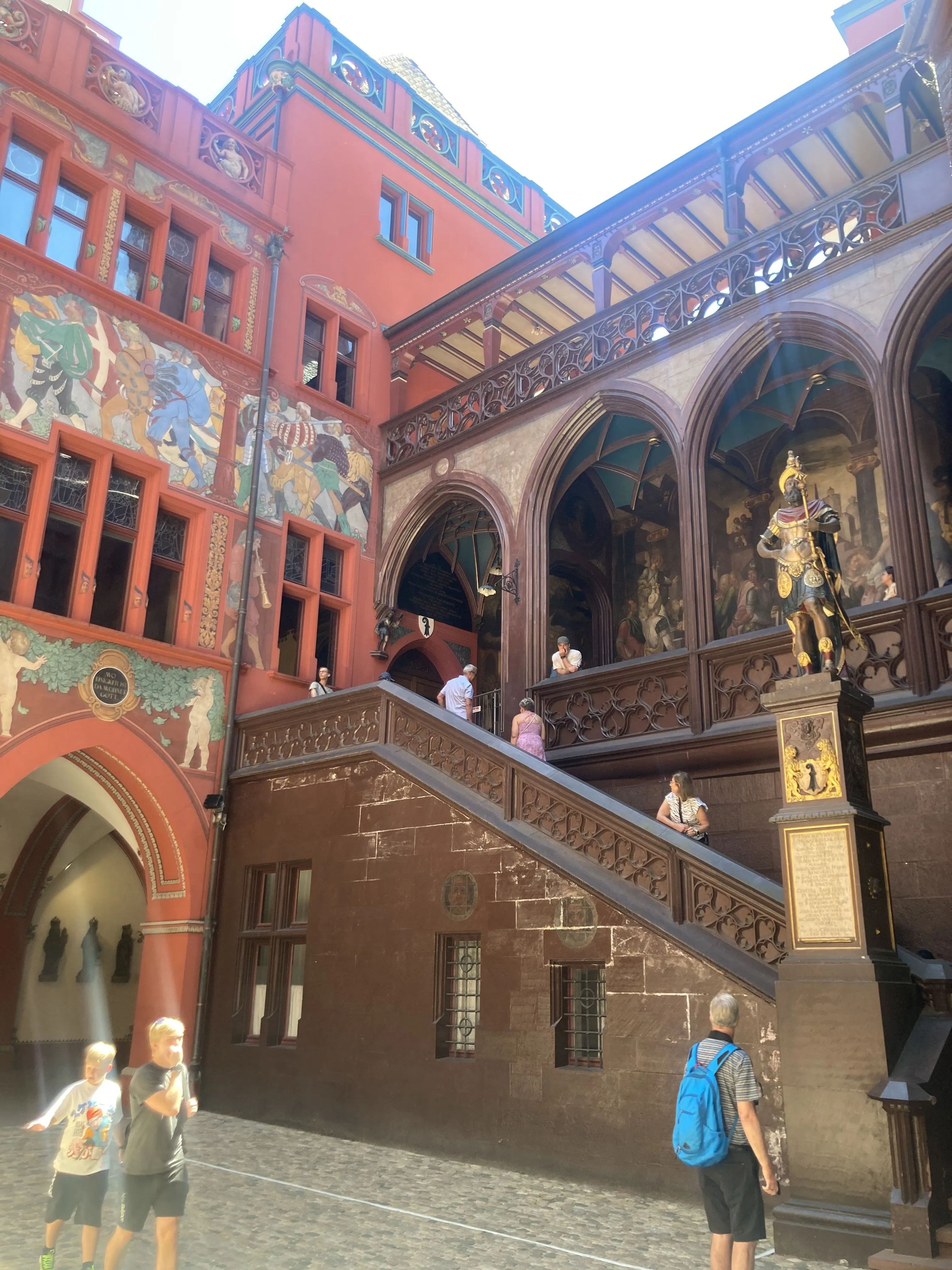 Binnenplaats van het stadhuis van Bazel met een rood geschilderde gevel vol fresco's, een sierlijke houten trap met gewelfde galerijen, een standbeeld op een voetstuk en bezoekers die over de kasseien lopen
