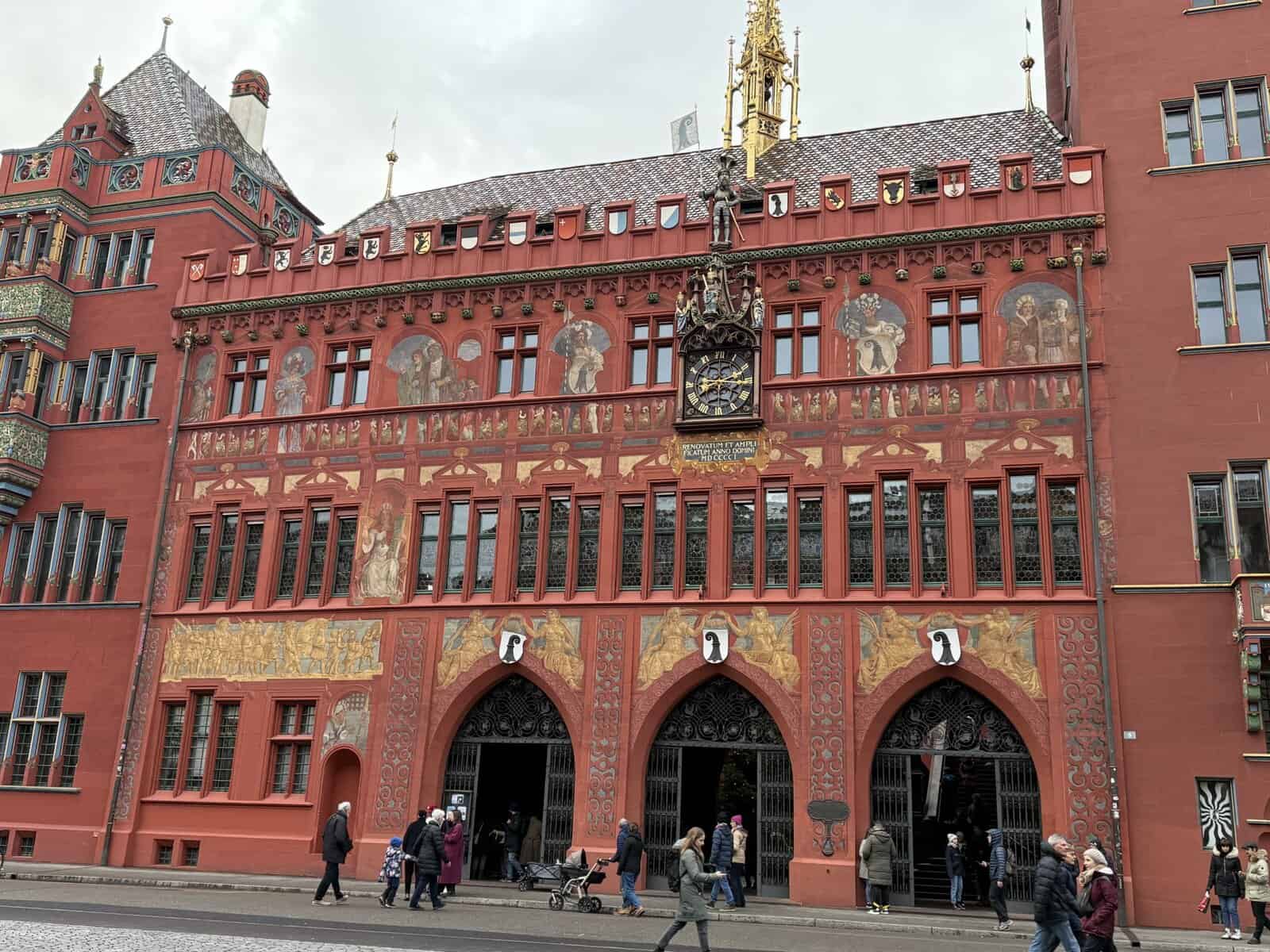 Rode gevel van het stadhuis van Bazel met beschilderde muurschilderingen, boogingangen en een gouden torenspits op het dak, met mensen die ervoor lopen