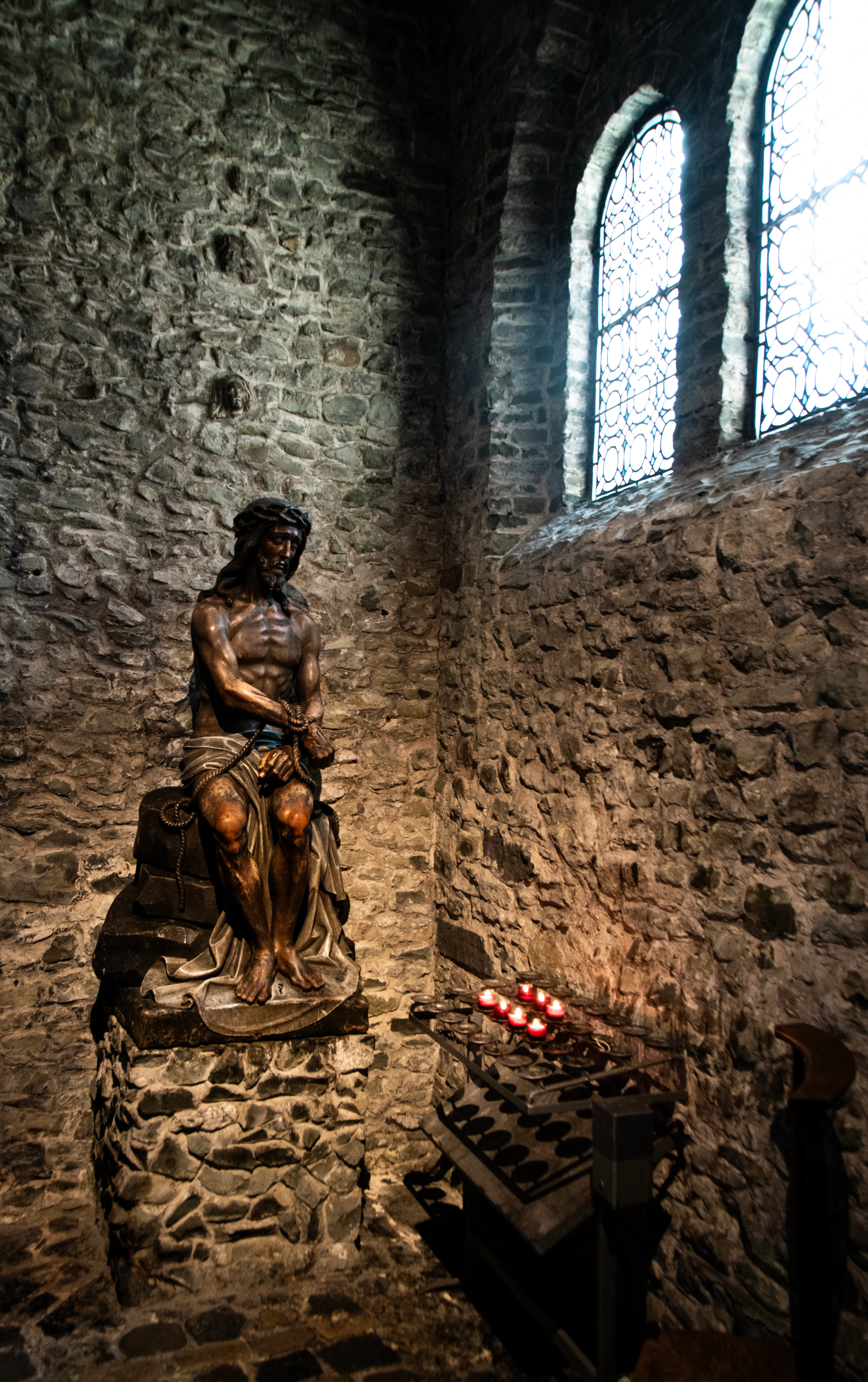A gilded wooden sculpture of a seated Jesus wearing a crown of thorns, displayed on a stone pedestal inside a medieval stone chapel with arched windows and lit votive candles.