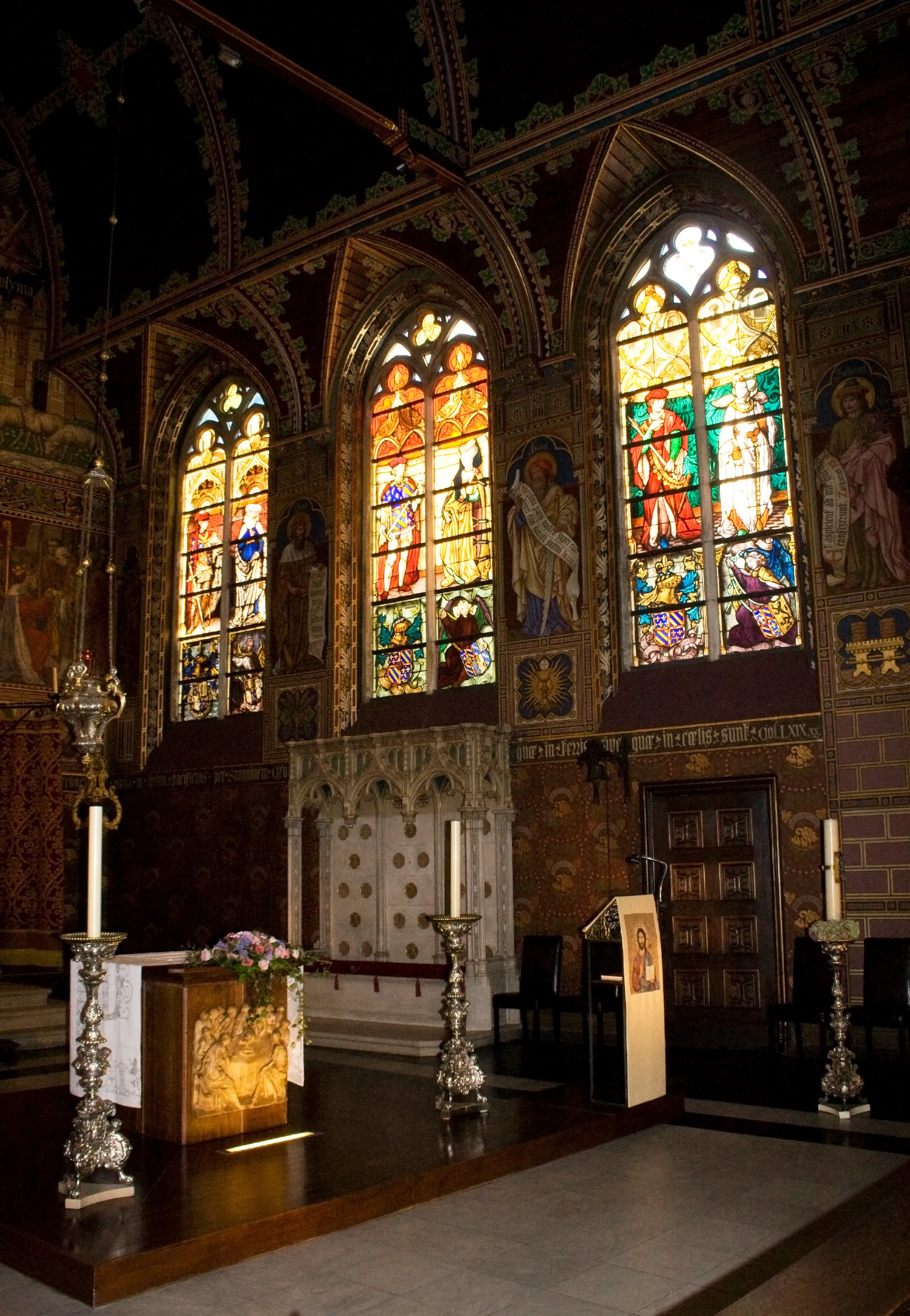 Interior of the Basilica of the Holy Blood showing the main nave with three large stained glass windows, a marble altar with silver candlesticks, and painted murals on the walls beneath Gothic arches.