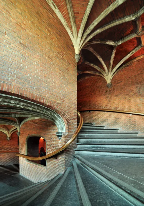 Spiral stone staircase with wooden handrail ascending through a brick vaulted chamber with ribbed Gothic ceiling