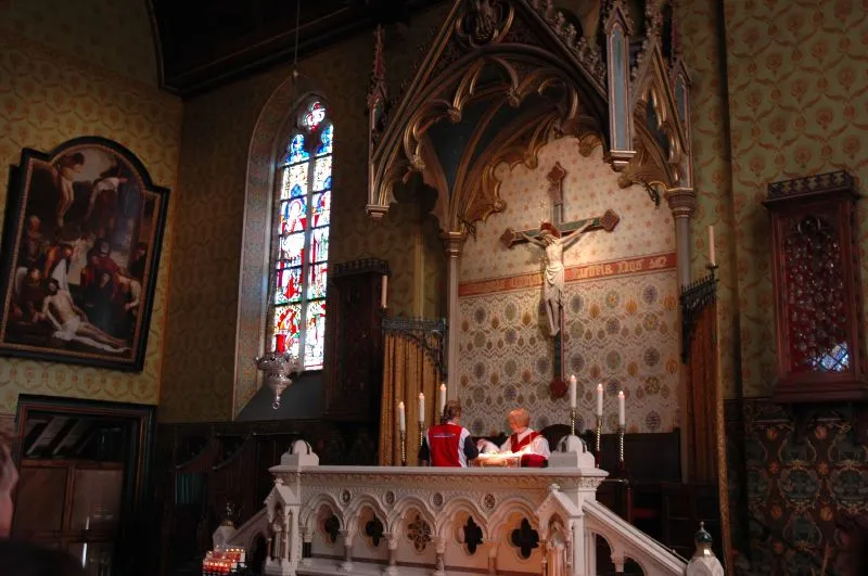 Interieur van de Heilig-Bloedbasiliek met een zijaltaar met een kruisbeeld, glas-in-loodraam en een priester in rode gewaden die een dienst leidt.