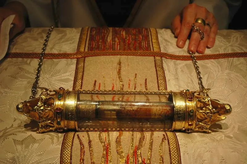 Rijkversierd gouden cilindervormig reliekhouder met decoratieve eindkappen rustend op een geborduurd wit en rood kleed, waarbij handen de bevestigde kettingen vasthouden