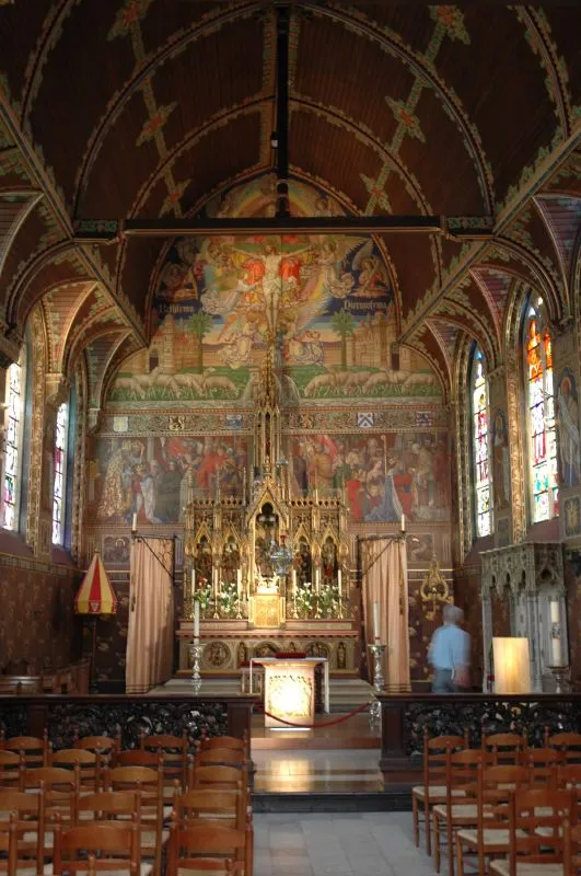 Interior of the Basilica of the Holy Blood showing the ornate main altar with Gothic detailing, colorful murals covering the apse walls, stained glass windows, and rows of wooden chairs