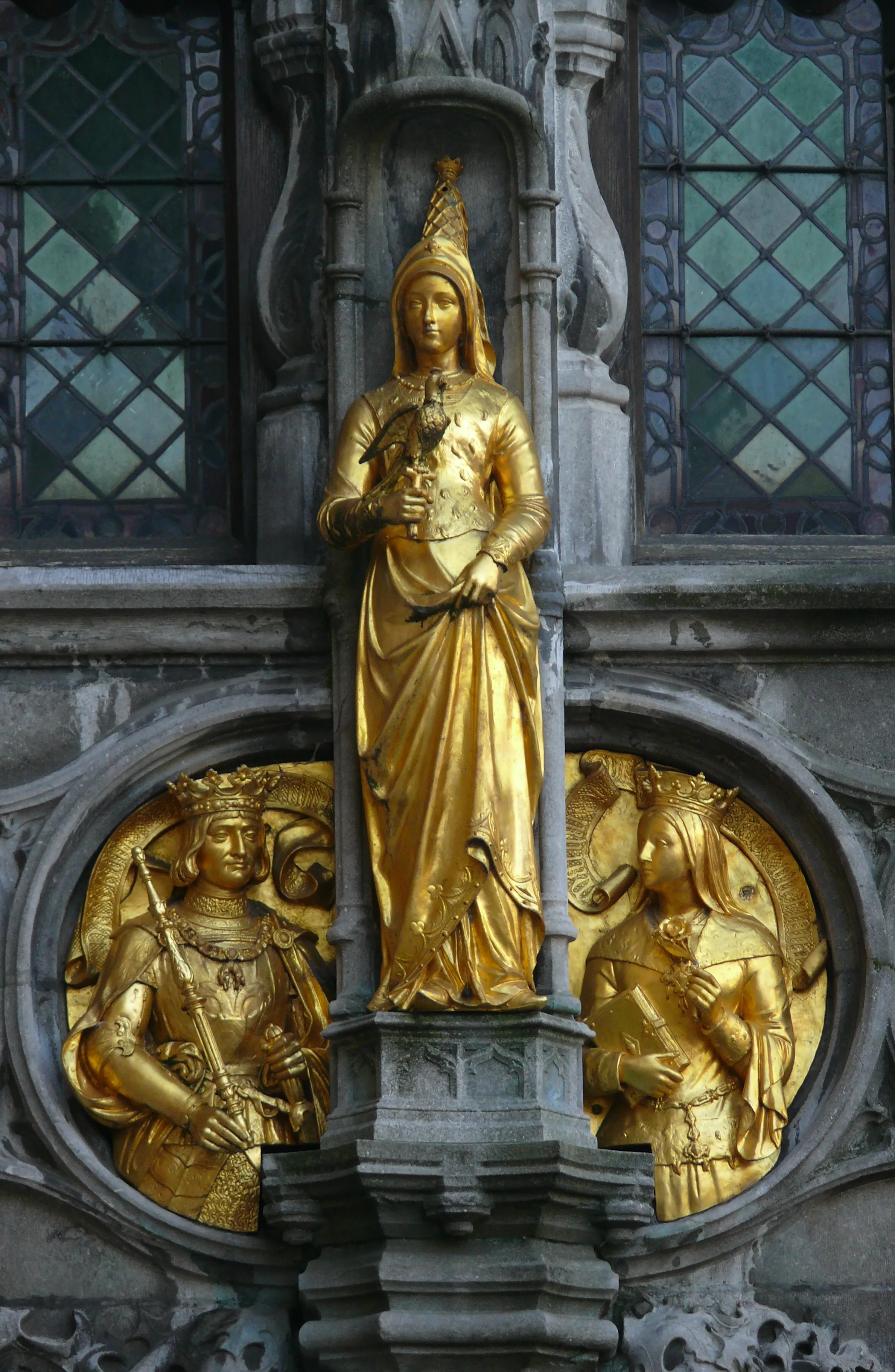 Golden statue of a crowned woman standing in a Gothic stone niche above two circular golden reliefs of crowned figures on the basilica facade, with stained glass windows visible behind.