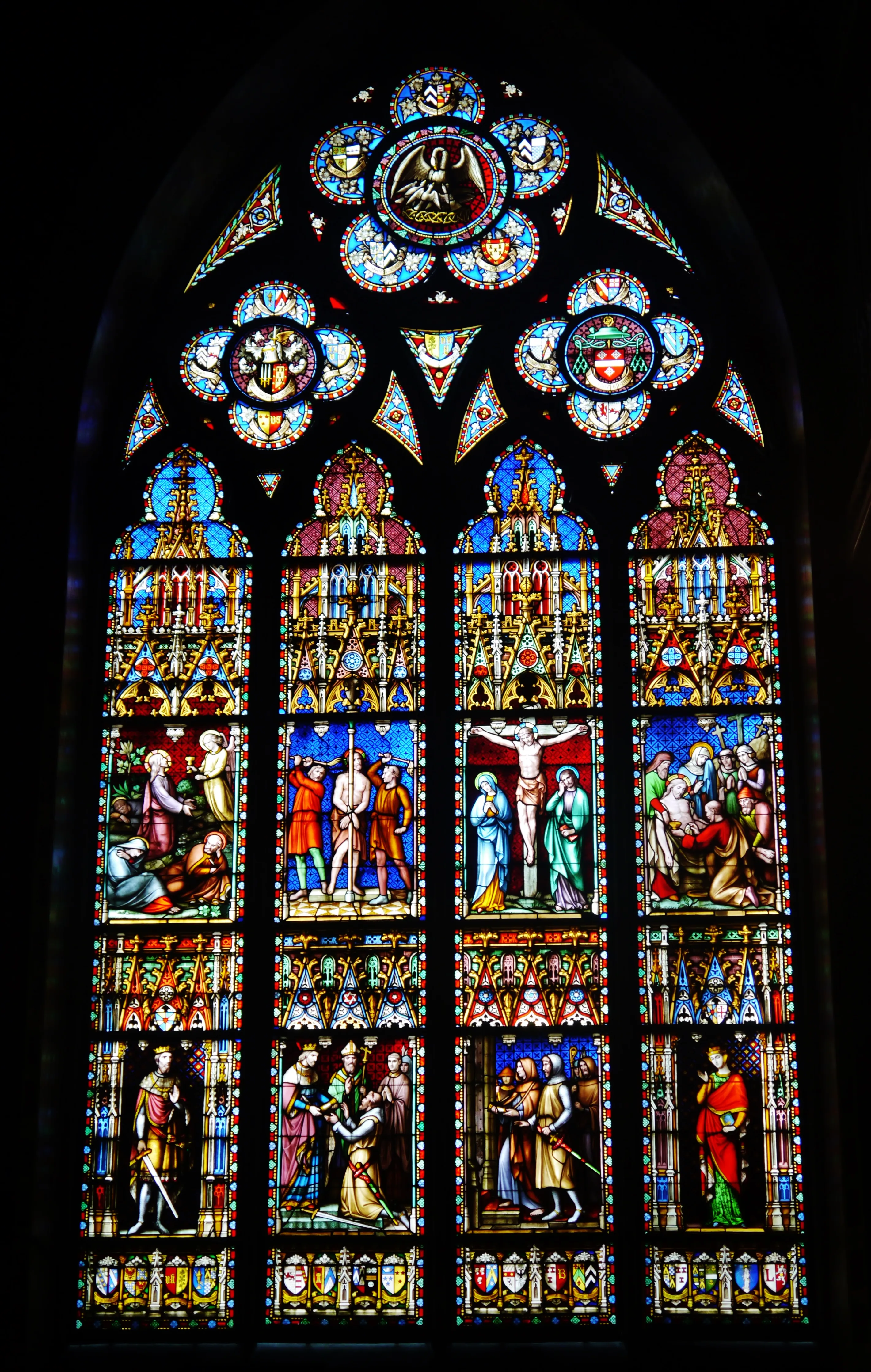 Tall arched stained glass window featuring colorful panels of biblical scenes including the crucifixion and heraldic crests at the top against a dark background.