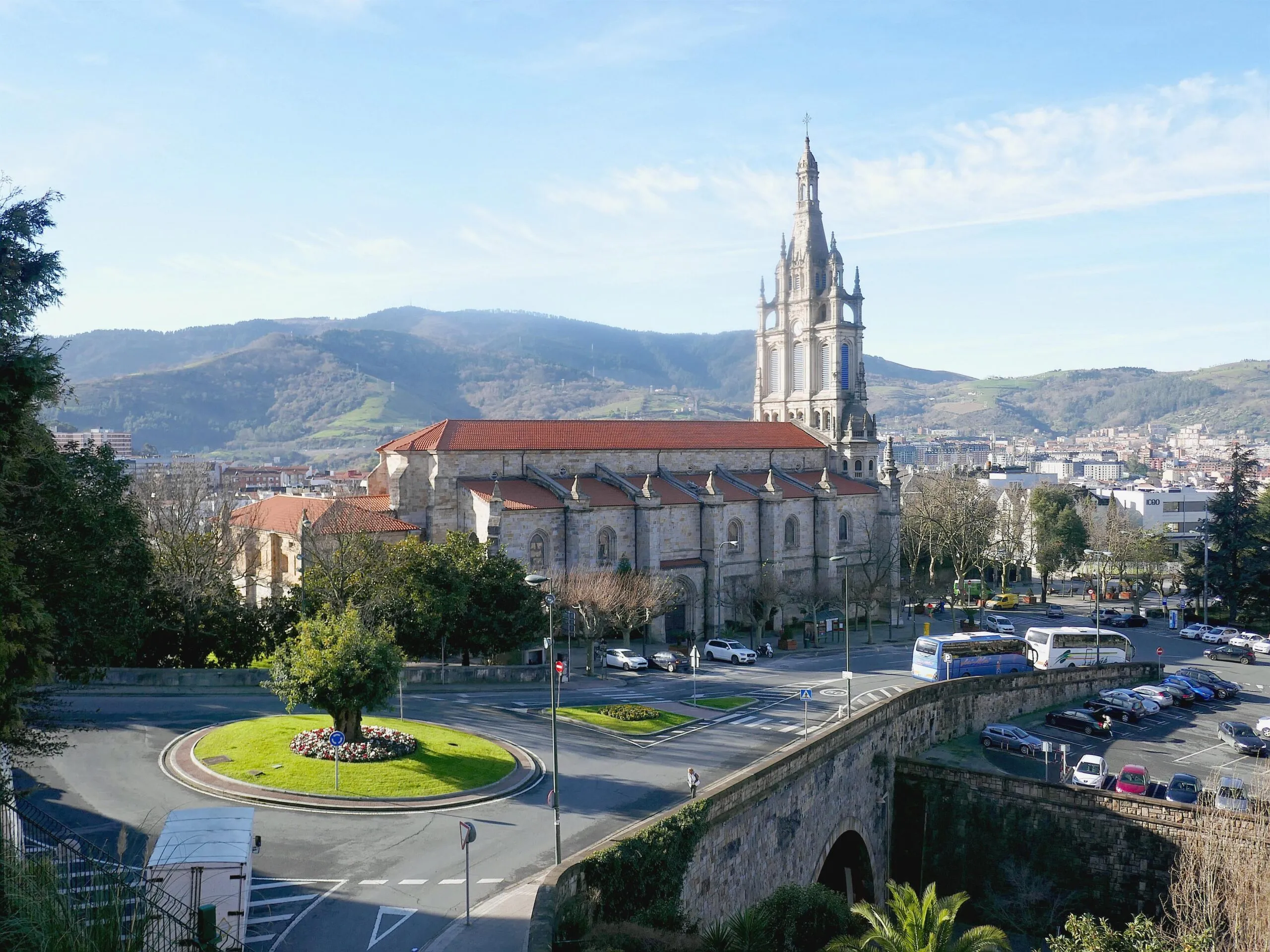 Widok z zewnątrz na Basilica of Begoña ukazujący jej wysoką gotycką wieżę, dach z czerwonej dachówki oraz otaczające rondo z górami w tle.