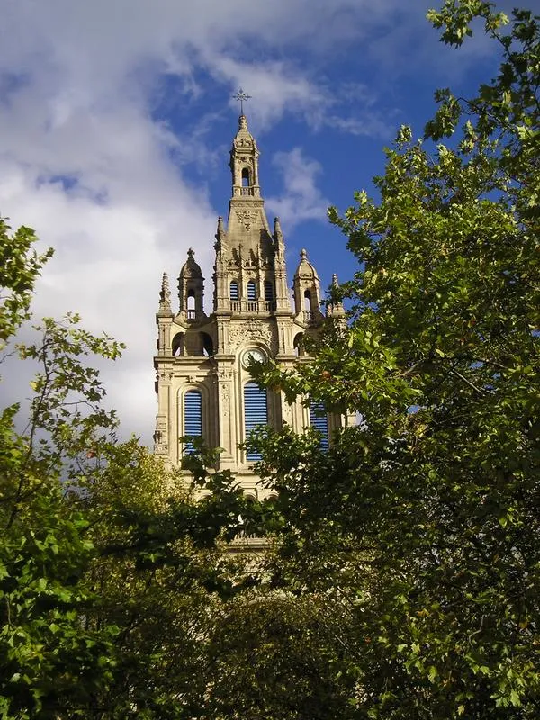 Ozdobna kamienna wieża bazyliki Basilica of Begoña wznosząca się ponad zielonymi drzewami na tle błękitnego nieba z białymi chmurami, zwieńczona krzyżem.