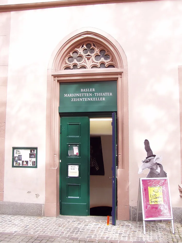 Zielone drzwi wejściowe do Basler Marionetten-Theater Zehntenkeller pod gotyckim oknem łukowym z kamiennym maswerkiem, z wyciętą postacią marionetki i szyldem po prawej stronie