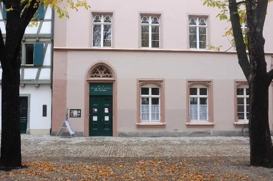 Różowa fasada budynku z zielonymi łukowymi drzwiami i oknami w białych ramach, otoczona dwoma ciemnymi pniami drzew z jesiennymi liśćmi rozrzuconymi na brukowanym podłożu