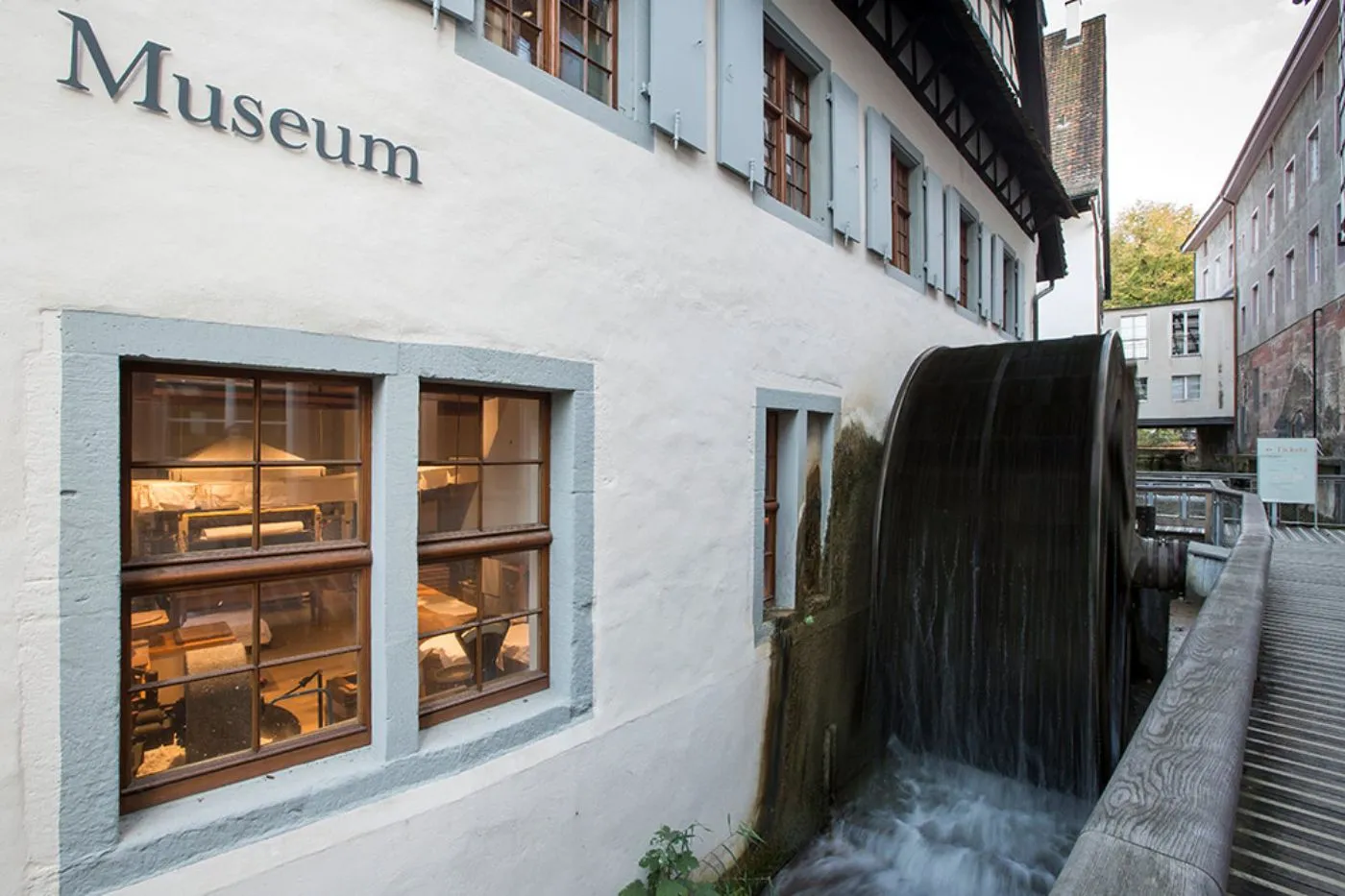 Fachada blanca de un edificio de museo con la palabra Museum en letras oscuras sobre ventanas con marco de madera, junto a una gran rueda de molino de madera oscura con agua fluyendo debajo.