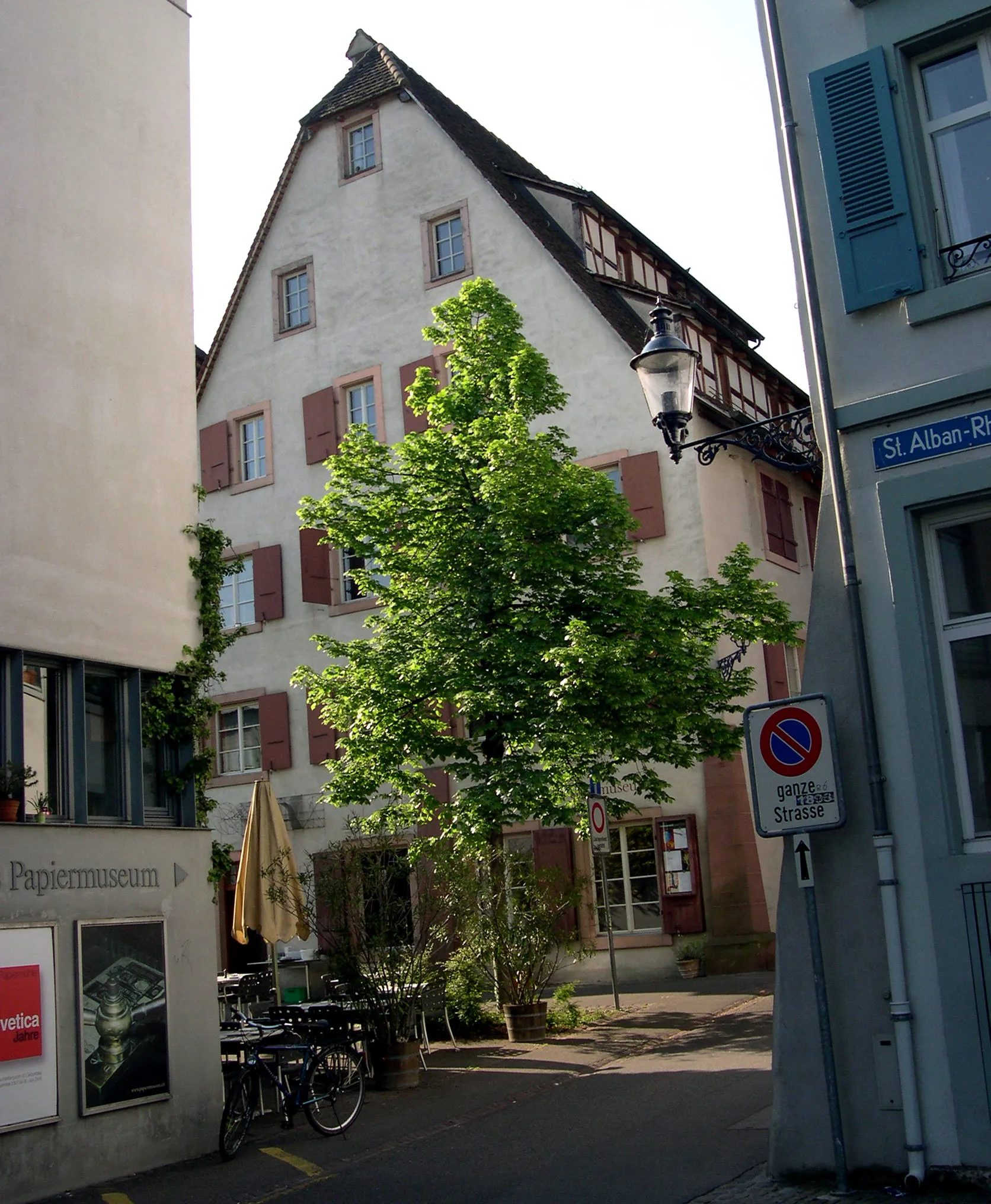 Vista de la calle del edificio de la Basler Papiermühle con fachada blanca, contraventanas rojas, un gran árbol verde, bicicletas aparcadas y un cartel de Papiermuseum en la pared adyacente