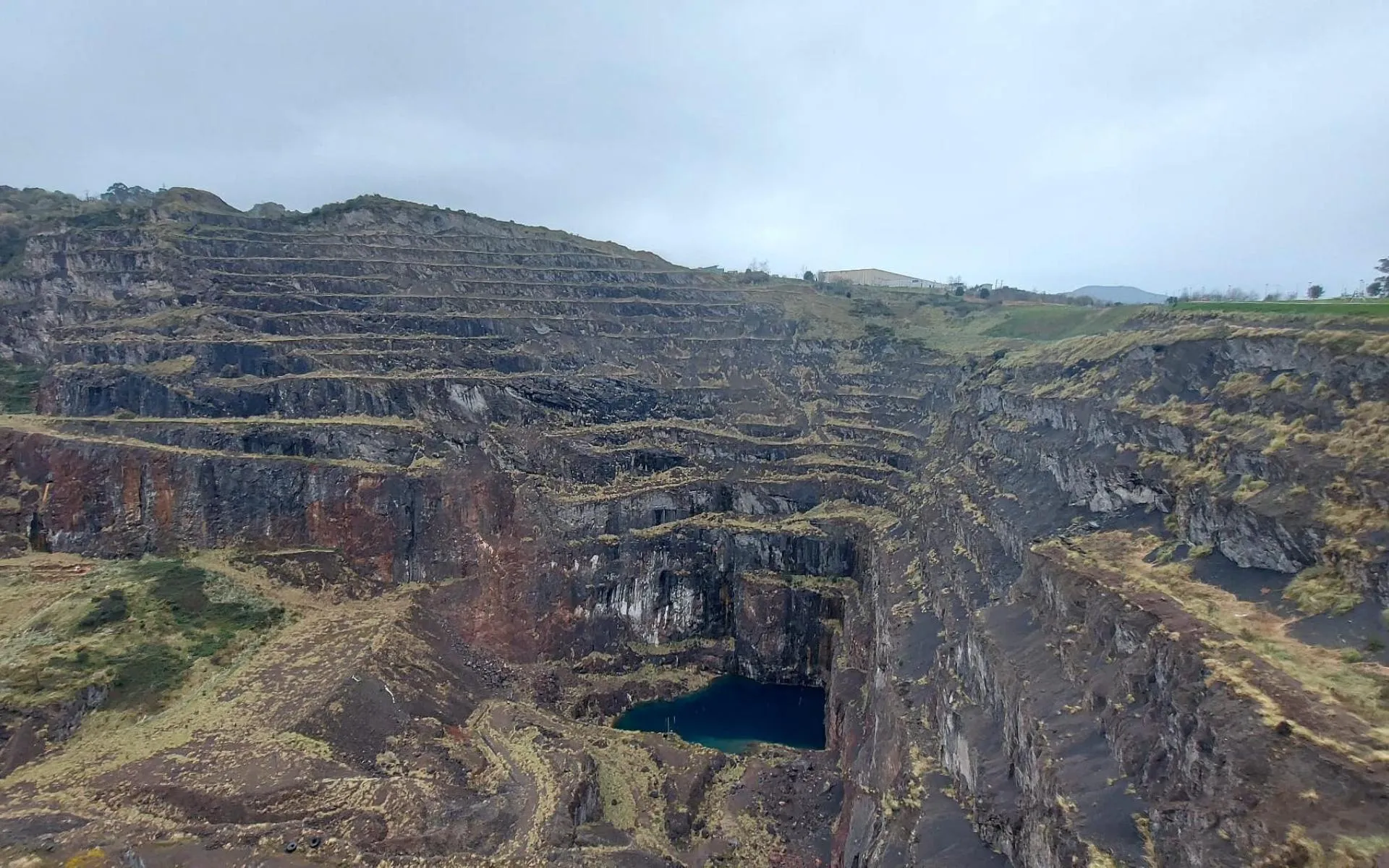 Vista panorámica de una mina a cielo abierto con terrazas, paredes rocosas escarpadas y una pequeña reserva de agua en el fondo bajo un cielo nublado.