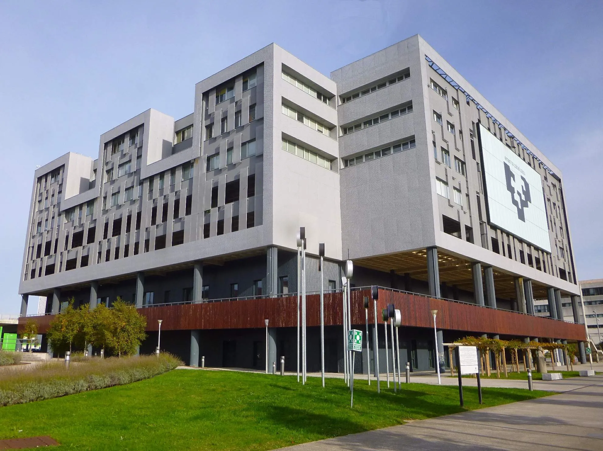 Large modern gray and white institutional building with a reddish-brown lower facade, set against a blue sky with green lawn in the foreground