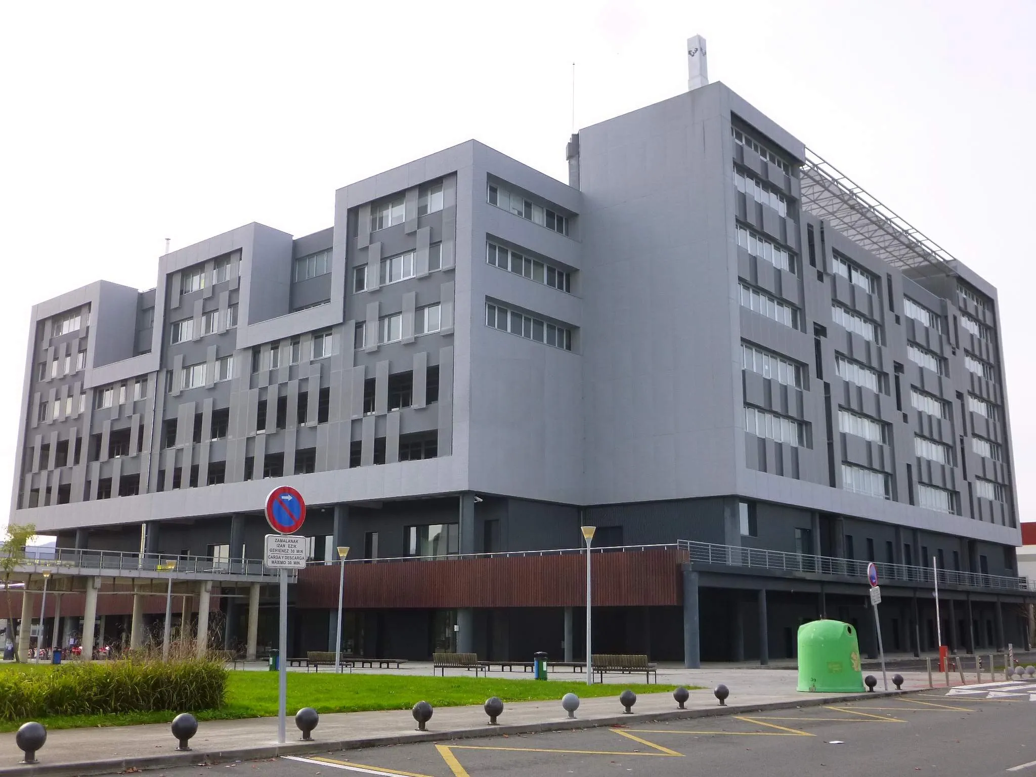 Large modern gray institutional building with stepped terraced facade, multiple stories of windows, and a brown lower section, viewed from a parking area with bollards and a green cylindrical structure in the foreground